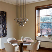 A split-view interior scene showing a dining room with a round wooden table, four white chairs, and a hanging chandelier, alongside a close-up of a gray dome pendant light against a wood panel and gray wall.